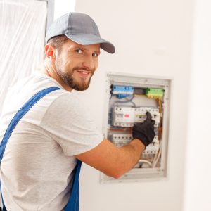 AdobeStock_484535932 Smiling electrician looking at camera while working with fuses at the circuit breaker control cabinet on the wall
